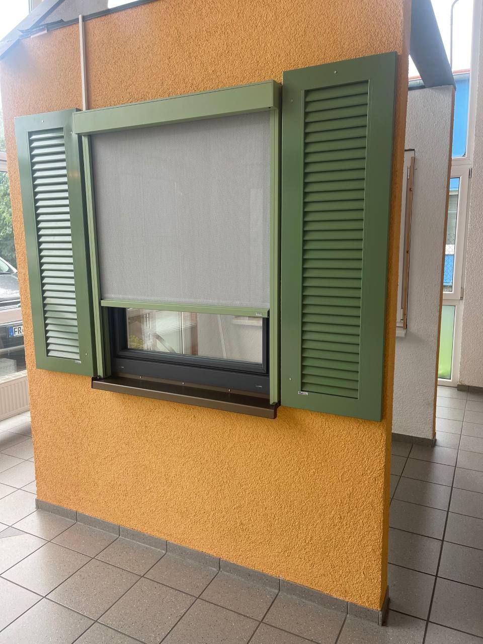 Eine gelbe, strukturierte Wand mit einem Fenster, das an beiden Seiten mit einem grauen Rollo und dekorativen grünen Fensterläden versehen ist.