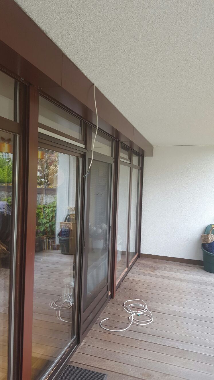 Eine Wohnterrasse mit großen Glasschiebetüren, Holzdielen und einer herumhängenden weißen Drahtrolle an der Decke.