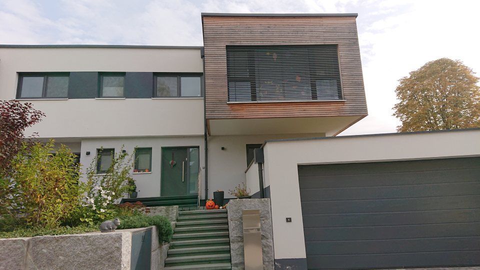 Ein modernes zweistöckiges Haus mit holzgetäfeltem Obergeschoss, weißer Fassade, dunklem Garagentor und einer Treppe im Eingangsbereich.