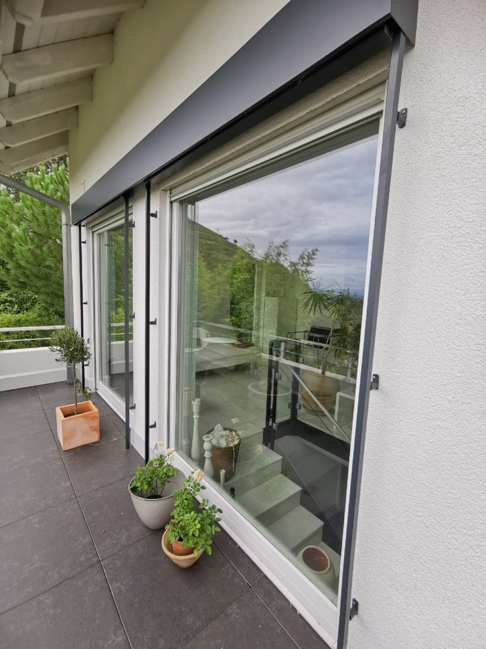 Ein hoher Balkon mit grauem Bodenbelag, ausgestattet mit zwei großen Glasschiebetüren mit schwarzen Rollladenkästen und -rahmen.