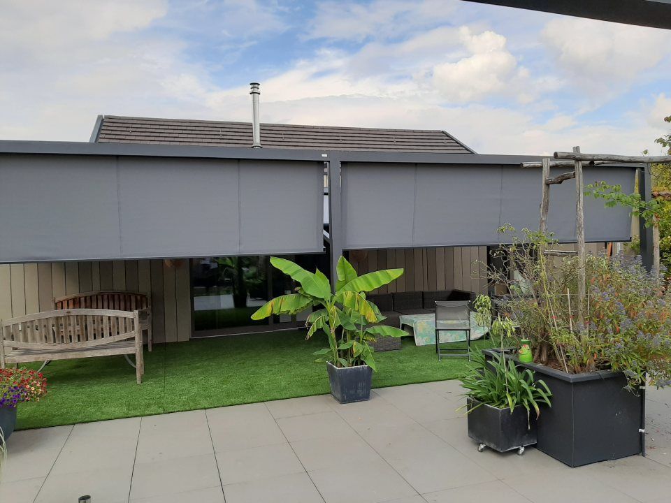 Ein Patio mit zwei grauen, herunterklappbaren Sonnenschutzdächern, einer Holzbank, Topfpflanzen und Kunstrasen unter einem Hausdach.