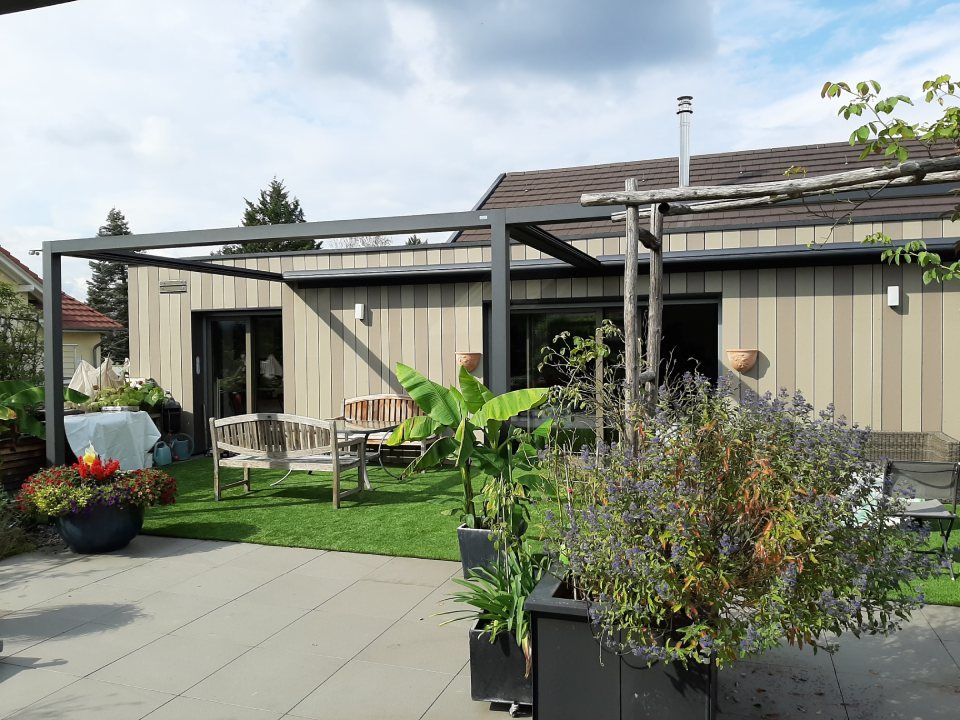 Eine moderne Terrasse mit Metallpergola, Kunstrasen, Gartenmöbeln und Topfpflanzen vor einem hellen Holzhaus.
