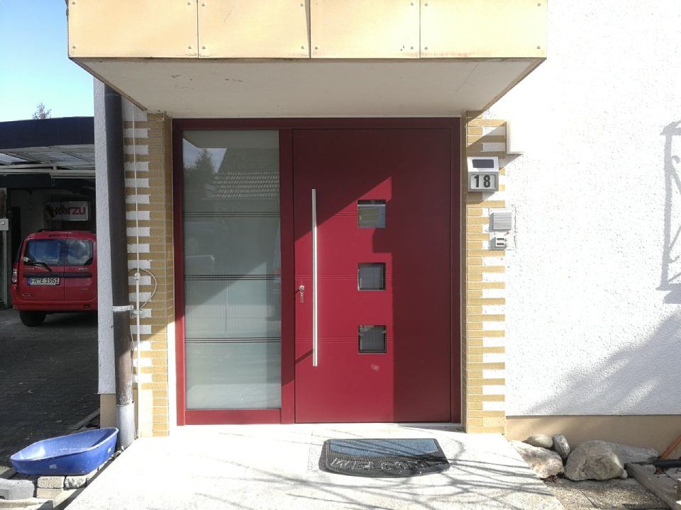 Eine moderne rote Haustür mit einem vertikalen silbernen Türgriff, einer seitlichen Glasscheibe, drei kleinen quadratischen Fenstern und einer Hausnummer.