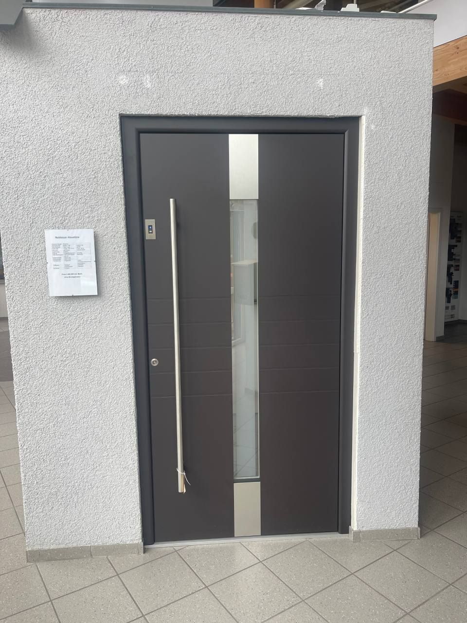 Eine dunkelgraue Haustür mit vertikaler Glasscheibe und langem silbernen Griff, eingelassen in eine strukturierte hellgraue Wand.