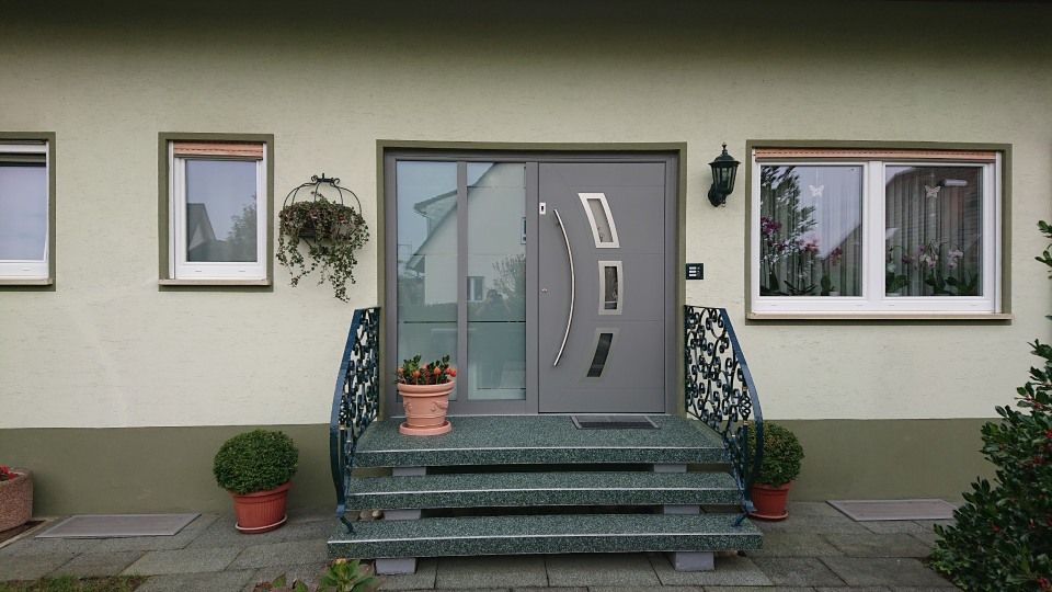 Eine moderne graue Haustür mit geschwungenem Griff und Seitenfenster, eingefasst zwischen Fenstern an einem hellen Haus mit Steintreppe.