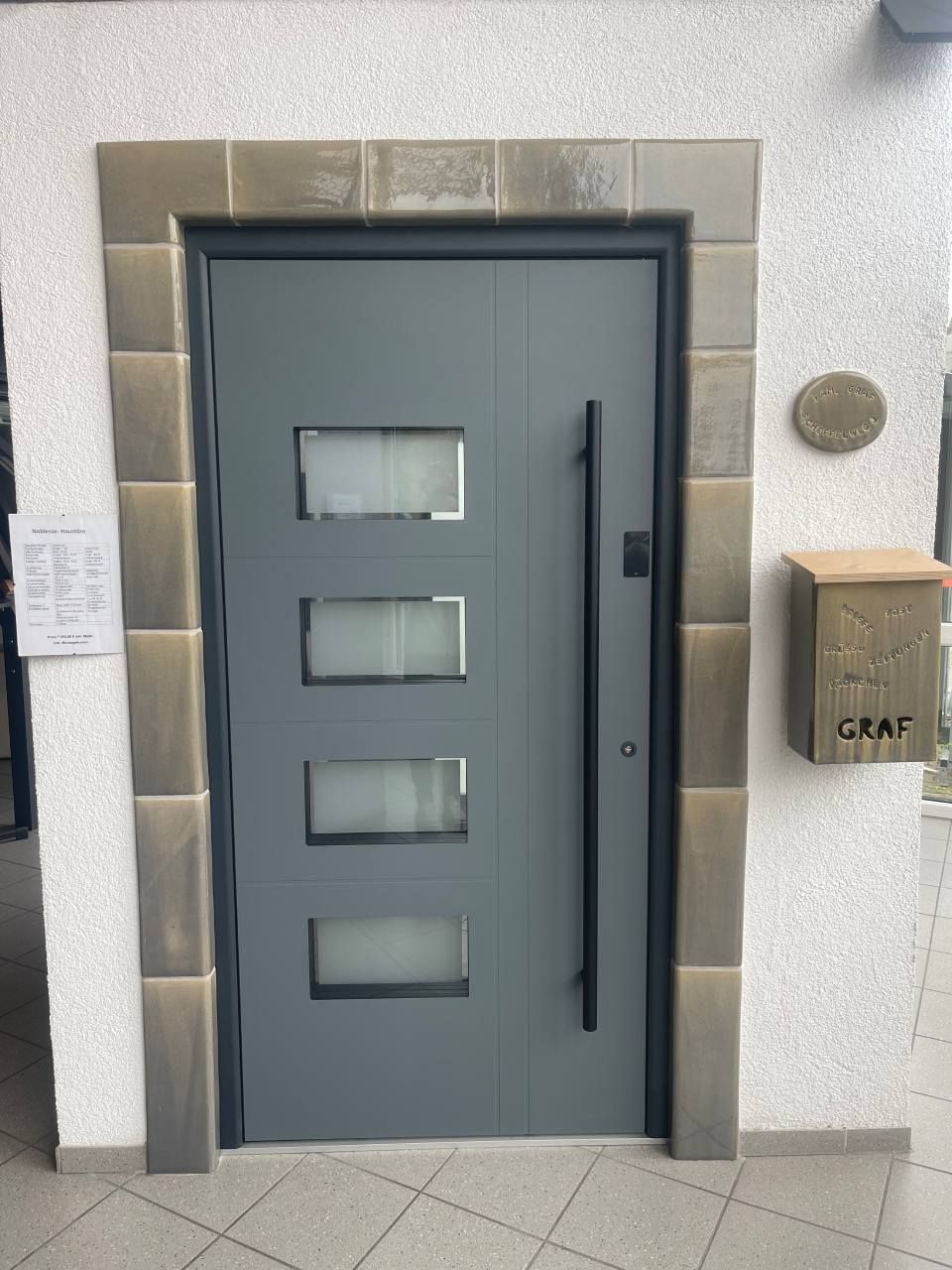 Eine graue, moderne Haustür mit vier rechteckigen Milchglasscheiben, einem hohen schwarzen Türgriff, Steinverzierungen und einem Wandbriefkasten.