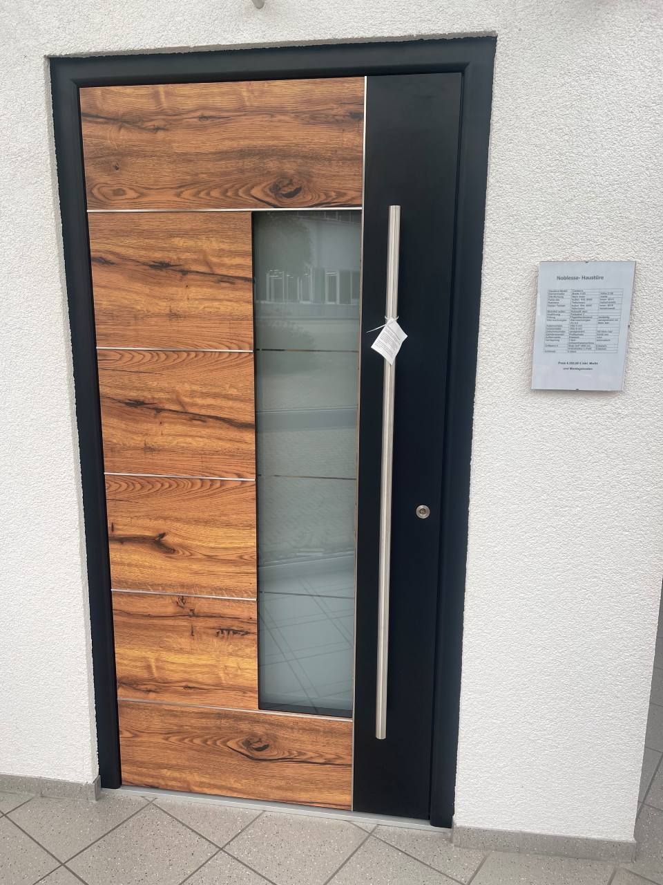 Eine moderne Tür mit Holzmaserung-Paneelen, einer vertikalen Milchglasscheibe und einem langen silbernen Griff in einem schwarzen Rahmen.