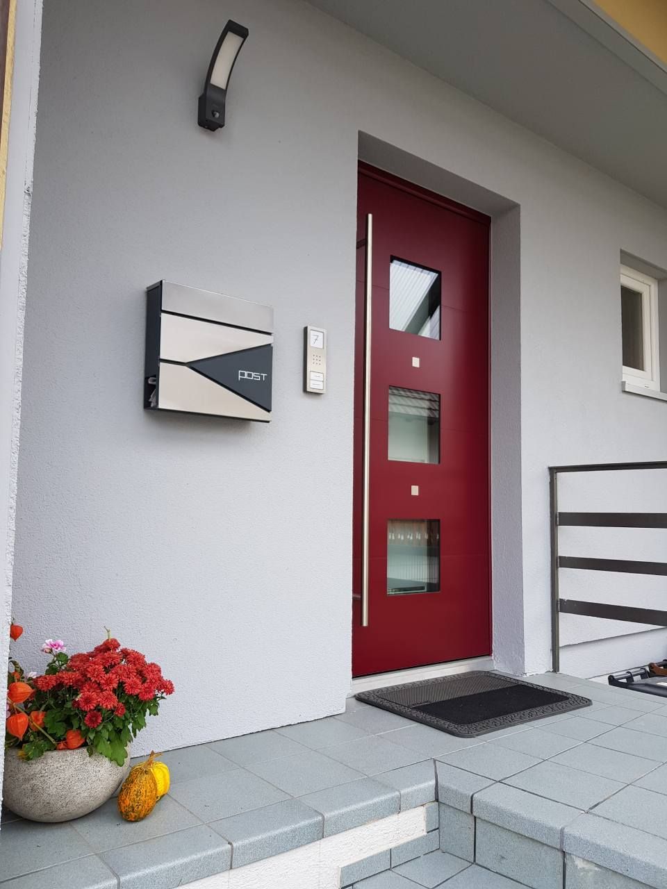 Eine rote Haustür an einem grauen Haus, dazu ein moderner Metallbriefkasten, eine Türklingel, eine Außenbeleuchtung und eine Topfpflanze.