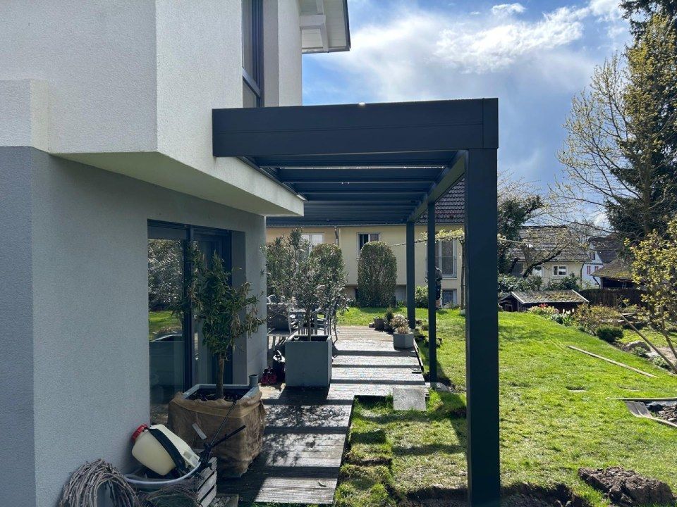 Moderne schwarze Metall-Terrassenpergola an einem hellen Haus mit Blick auf einen grasbewachsenen Hinterhof.