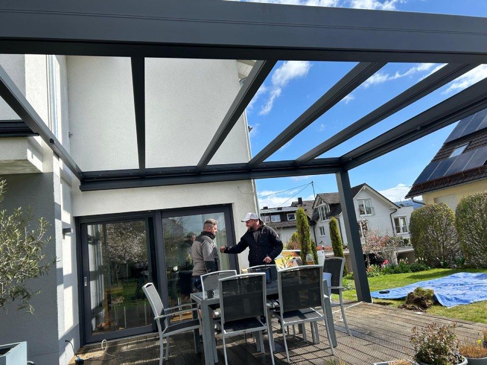 Zwei Personen stehen unter einer modernen, dunkelgrauen Terrassenpergola, die an ein weißes Haus angebaut ist, in der Nähe einer Essgruppe im Freien.