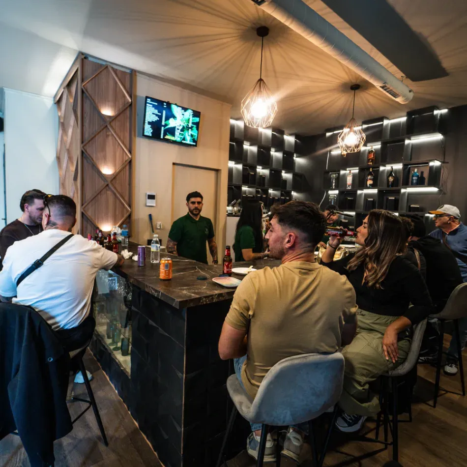 Gente sentada en un bar con poca luz, con un camarero y pantallas de televisión montadas en la pared.