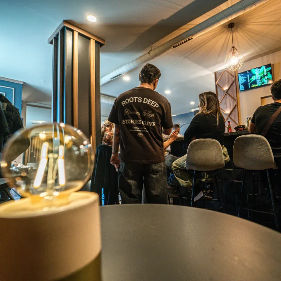 Cafetería moderna con iluminación tenue, gente sentada en la barra, lámparas colgantes cálidas y un televisor en la pared.