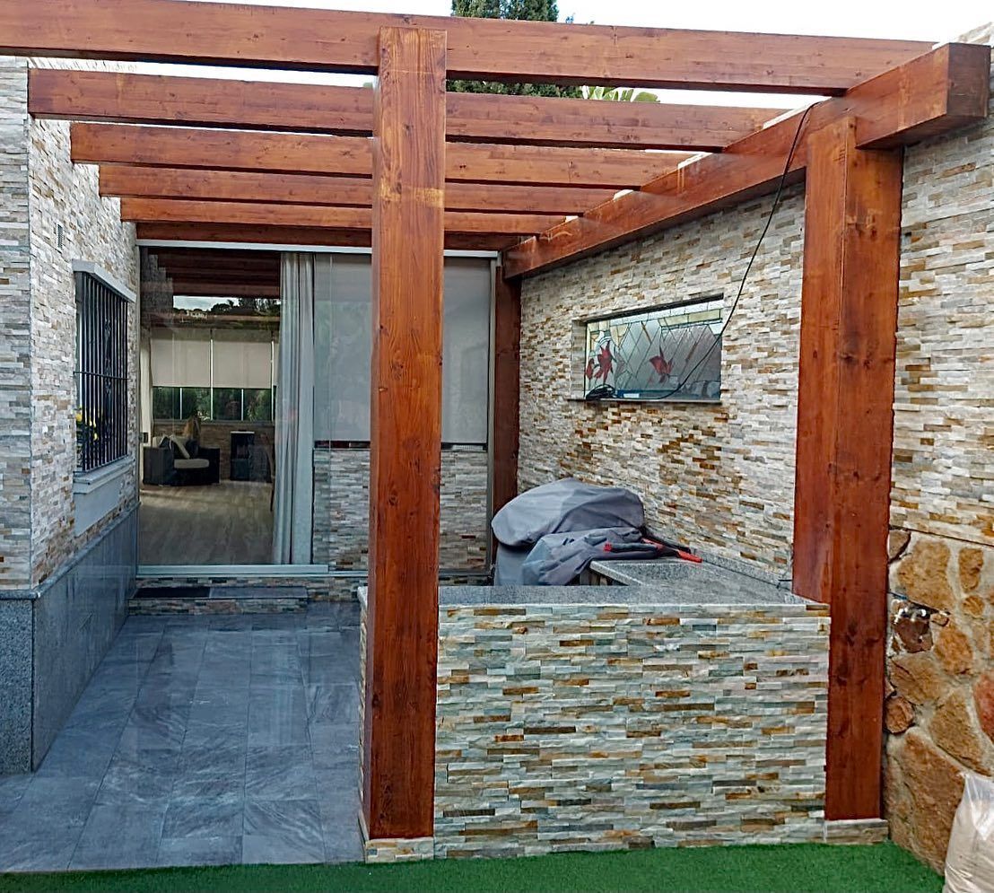 Una pérgola de madera está situada encima de un muro de piedra al lado de una casa.