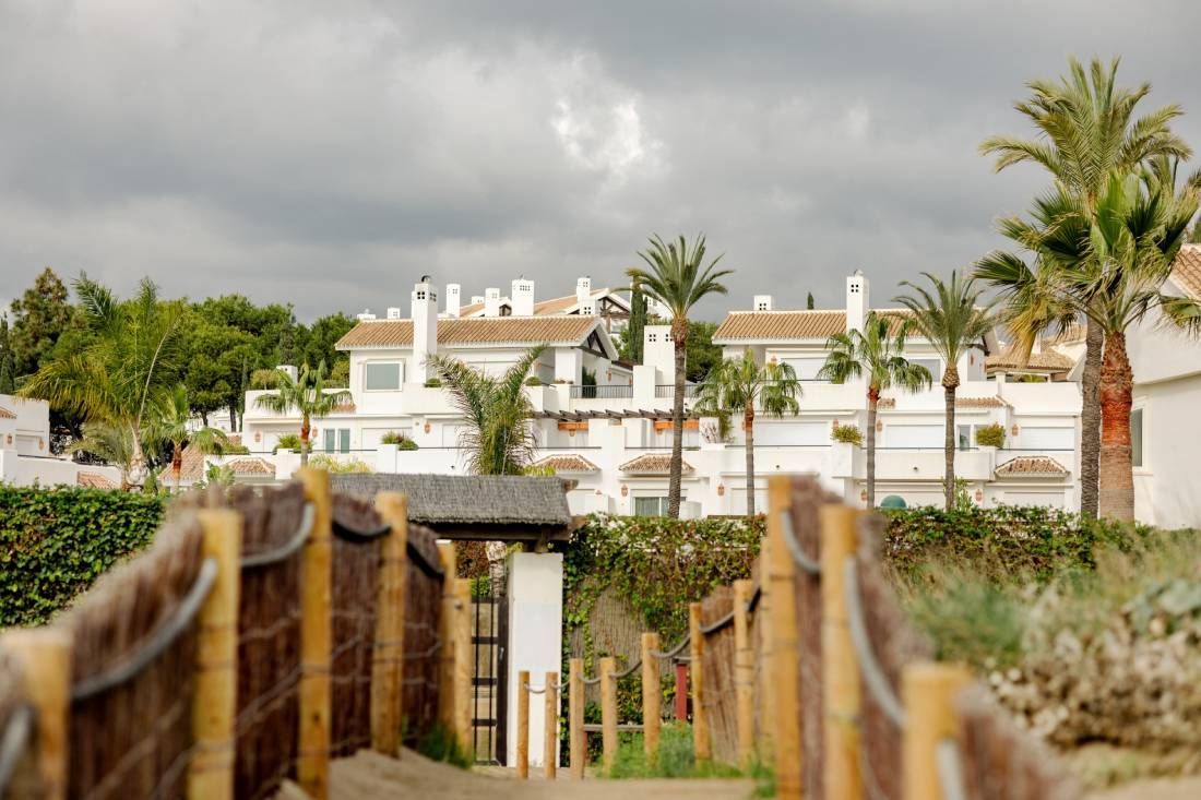 Una pasarela de madera que conduce a una casa blanca con palmeras al fondo.
