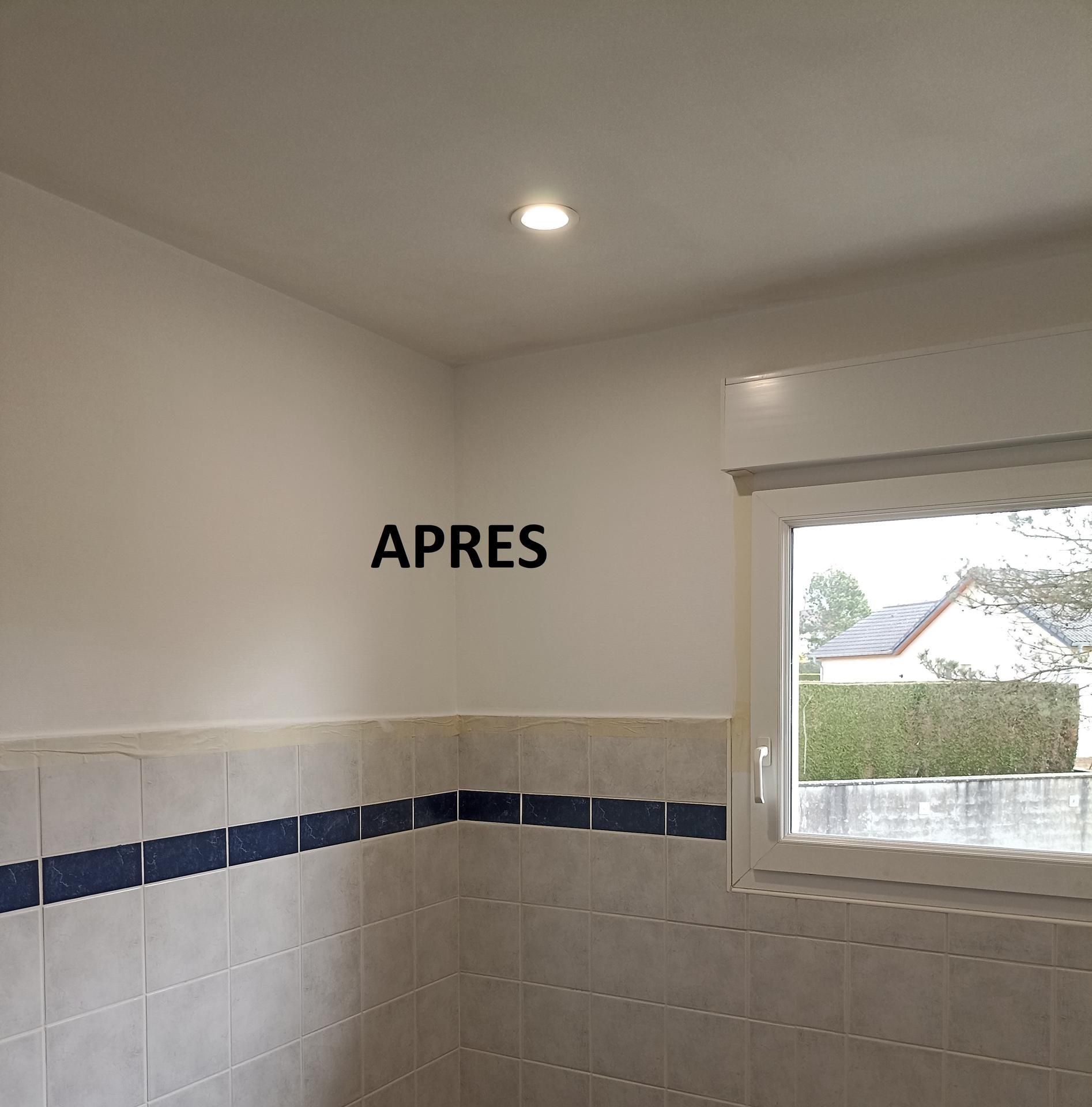 Coin de salle de bain avec murs carrelés blancs, bande décorative bleue, fenêtre et plafonnier ; inscription « APRES » sur le mur