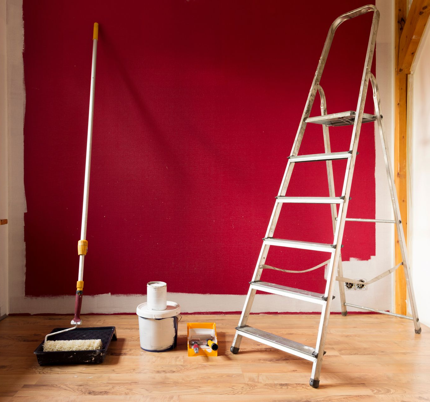 Mur peint en rouge avec matériel de peinture : rouleau, pot de peinture, bac à peinture, escabeau.