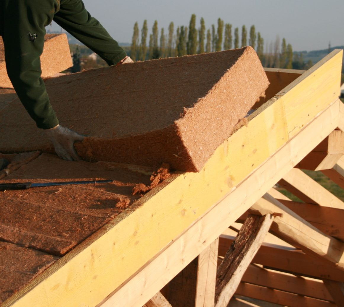 Une personne installe un panneau isolant brun sur une charpente de toit en bois.