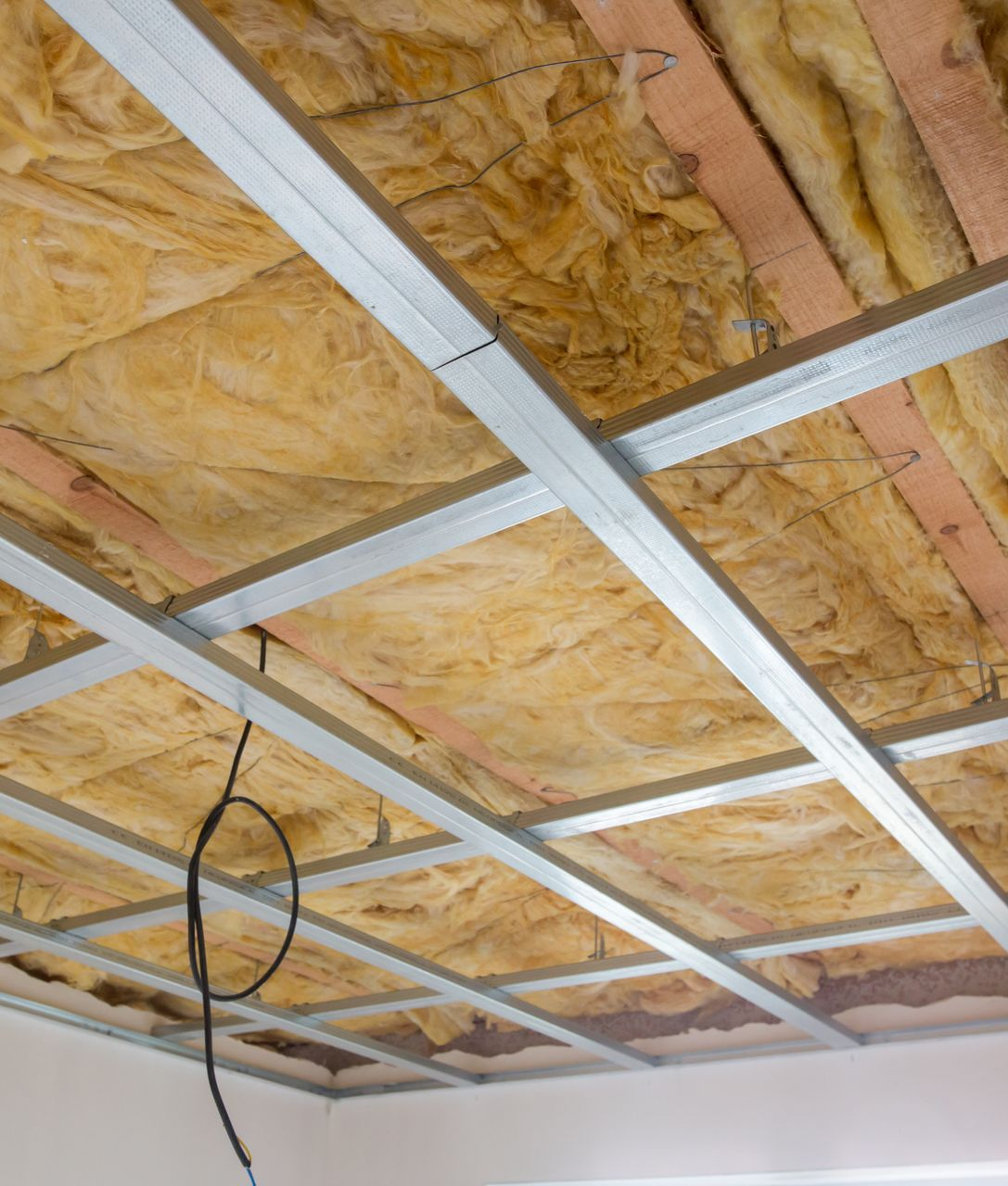 Isolation et grille métallique installées au plafond, avec des poutres en bois apparentes au-dessus et un fil électrique qui pend.