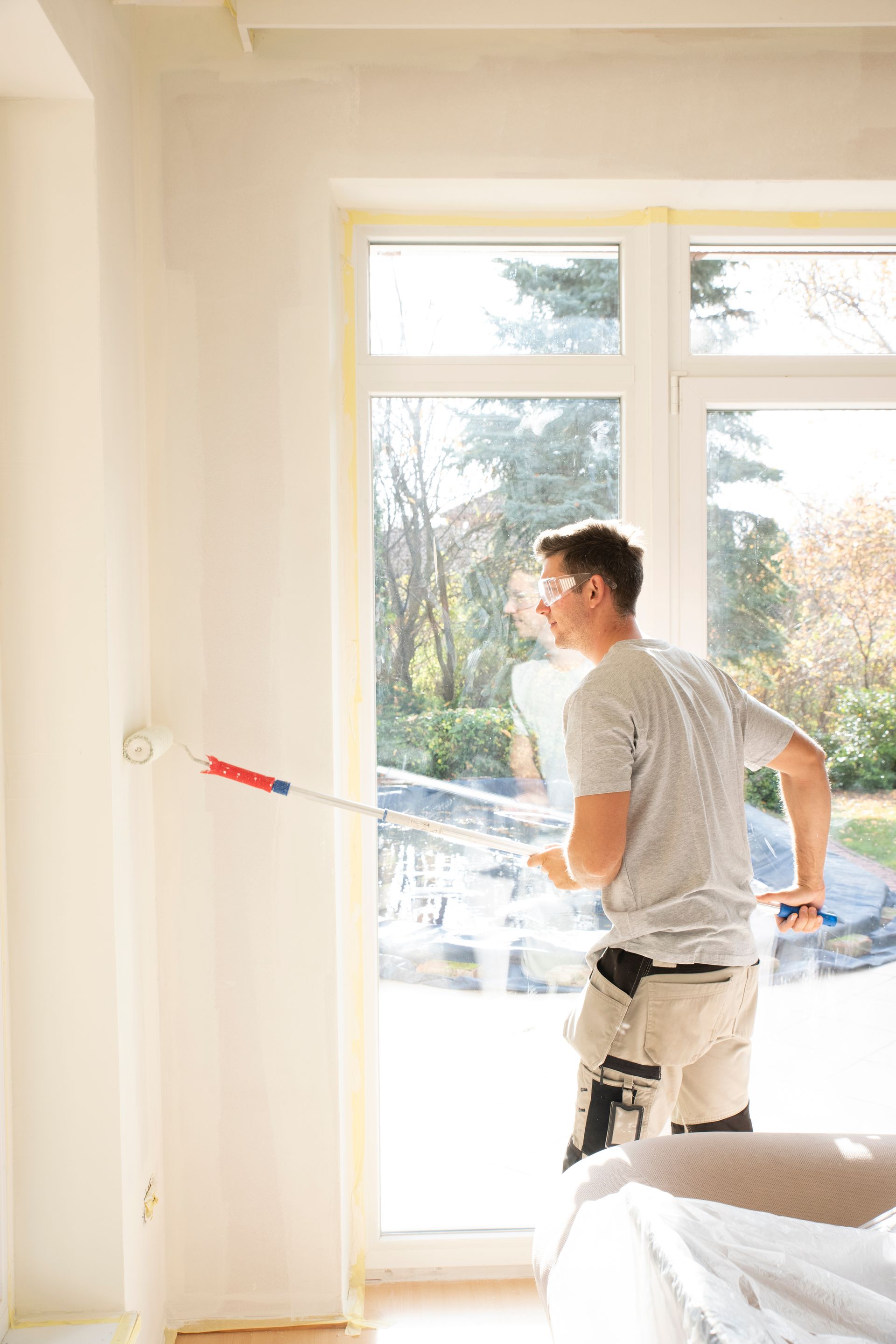 Un homme peint un mur au rouleau, à l'intérieur, près d'une fenêtre. Il semble sourire en travaillant.