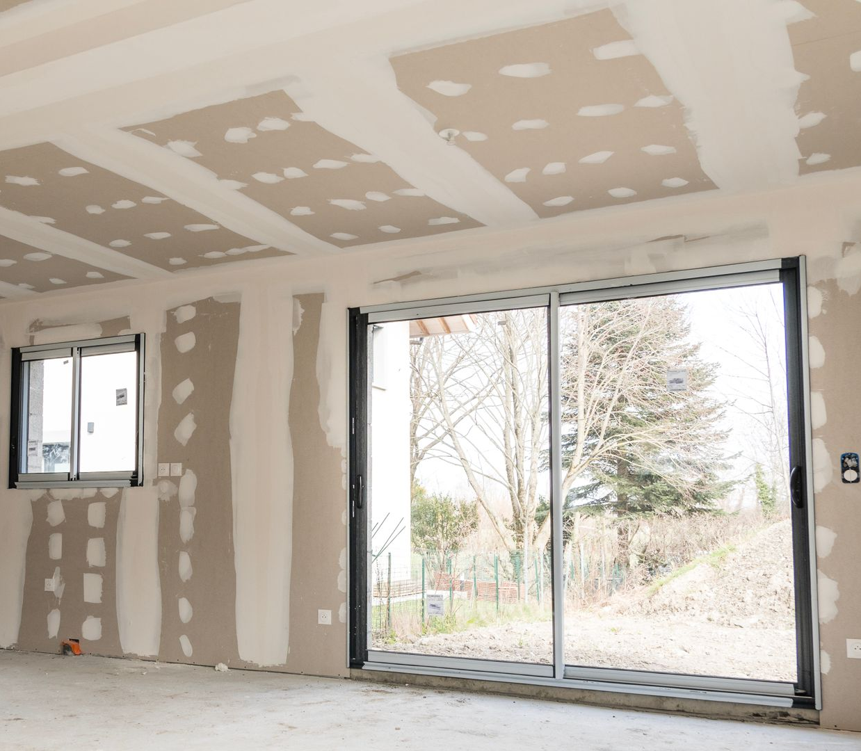 Intérieur en travaux, murs et plafond en plaques de plâtre. Grande et petite fenêtre visibles.