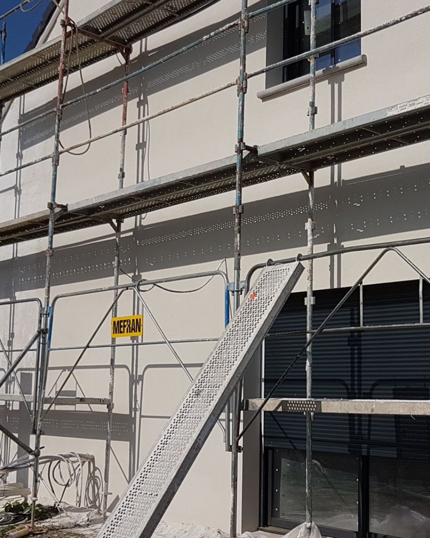 Échafaudages sur la façade d'un bâtiment ; des ouvriers effectuent probablement des travaux de rénovation extérieure. Murs clairs, échafaudages gris.