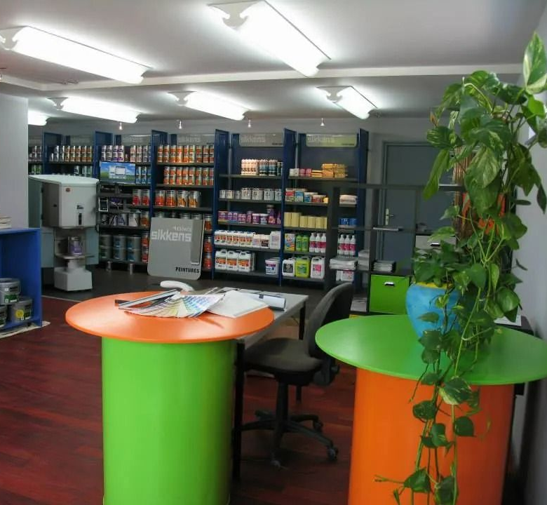 Intérieur d'un magasin de peinture avec des pots de peinture sur des étagères, deux tables colorées et une plante.
