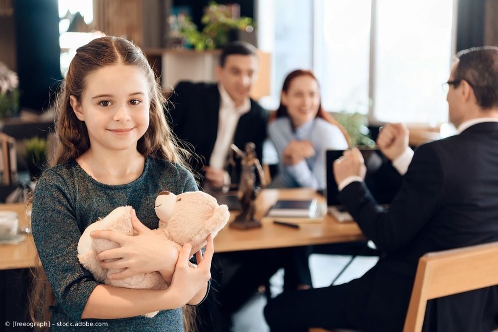 Ein Mädchen hält einen Teddybären in einem Bürobesprechungsraum, während Erwachsene im Hintergrund reden.