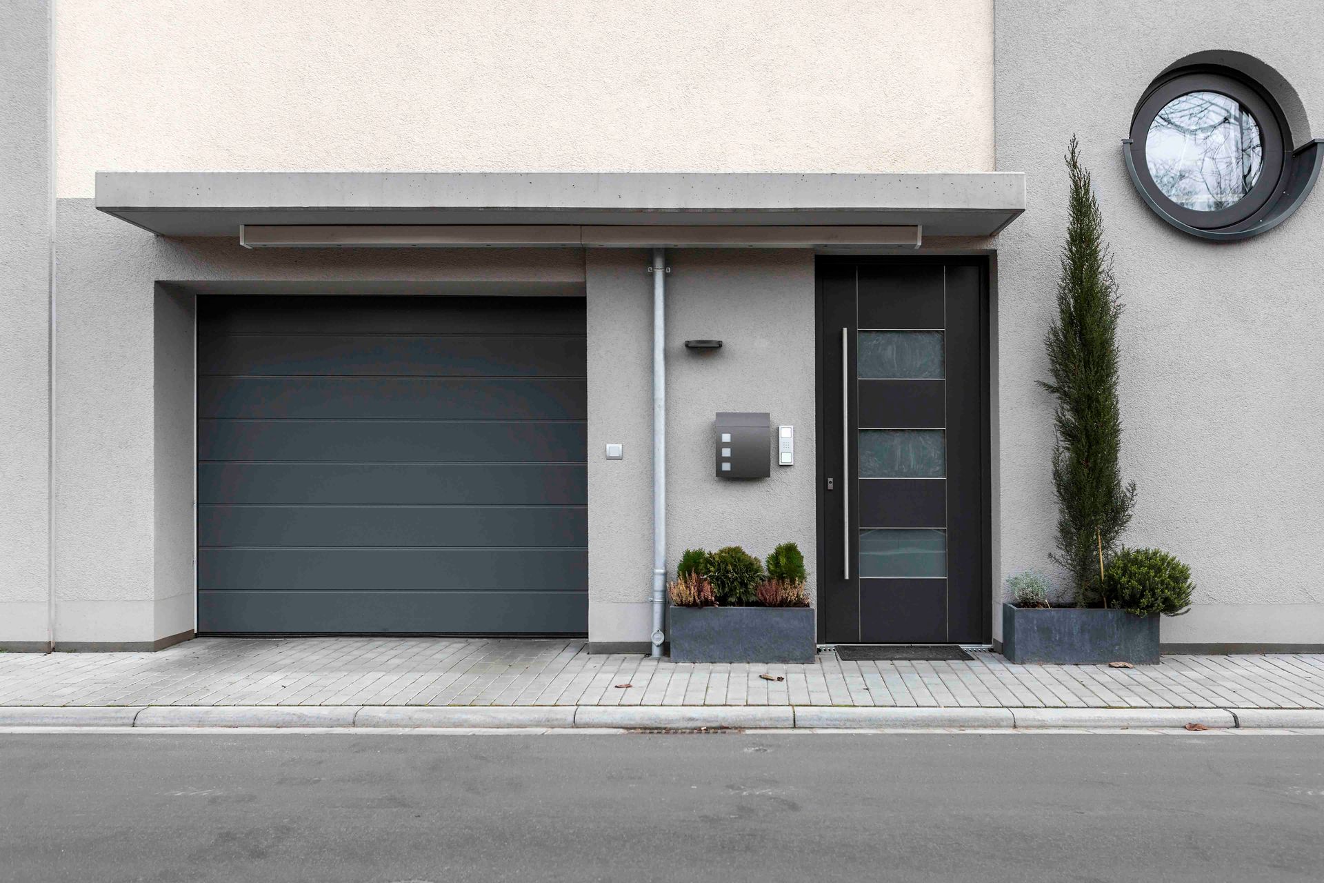 Moderne Hausfassade mit dunklem Garagentor, Glaseingang, Topfpflanzen und einer Wanduhr über der Tür.