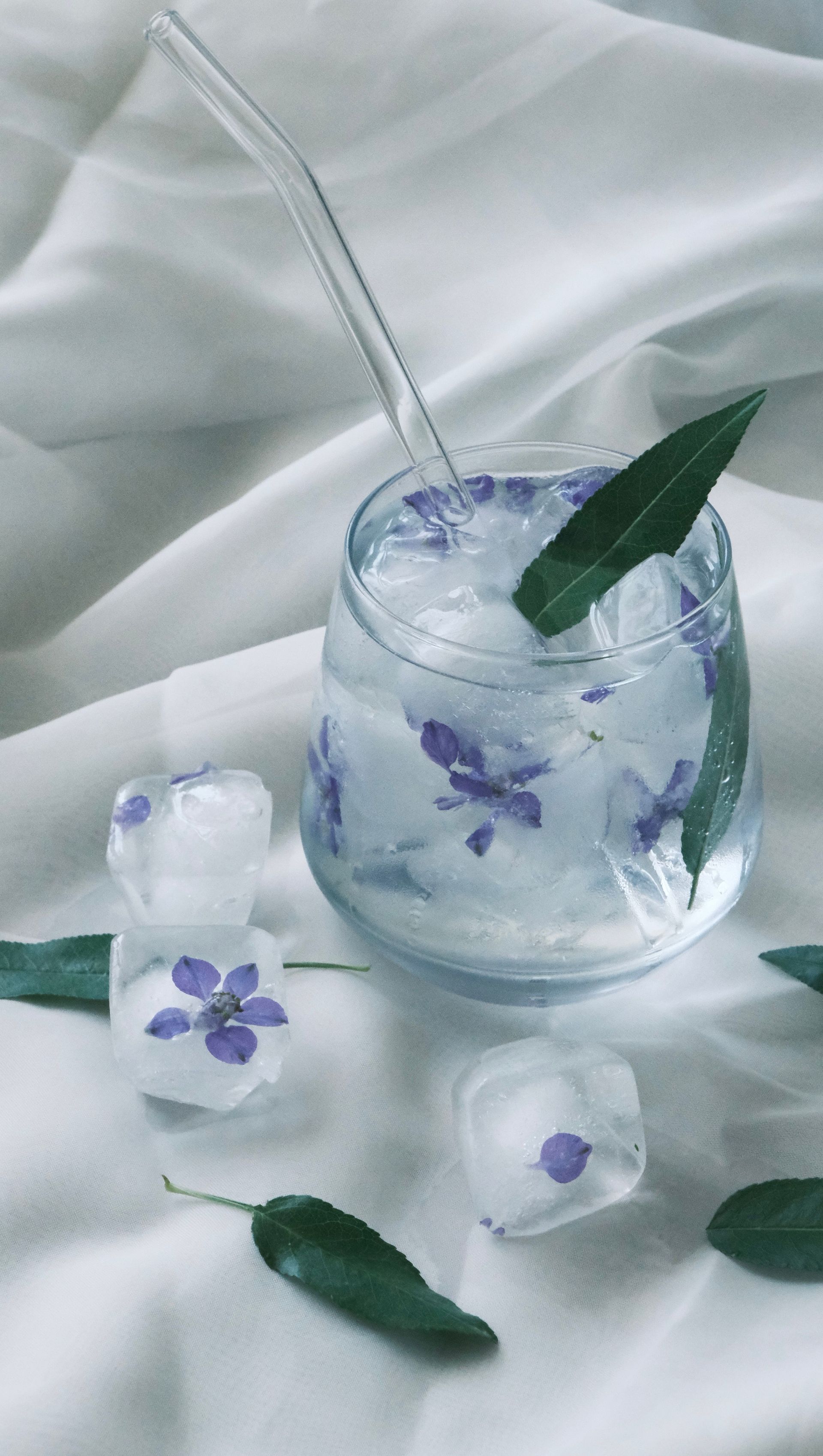 Boisson florale bleu glacé dans un verre transparent avec des feuilles de menthe, entourée de glaçons sur un tissu blanc.