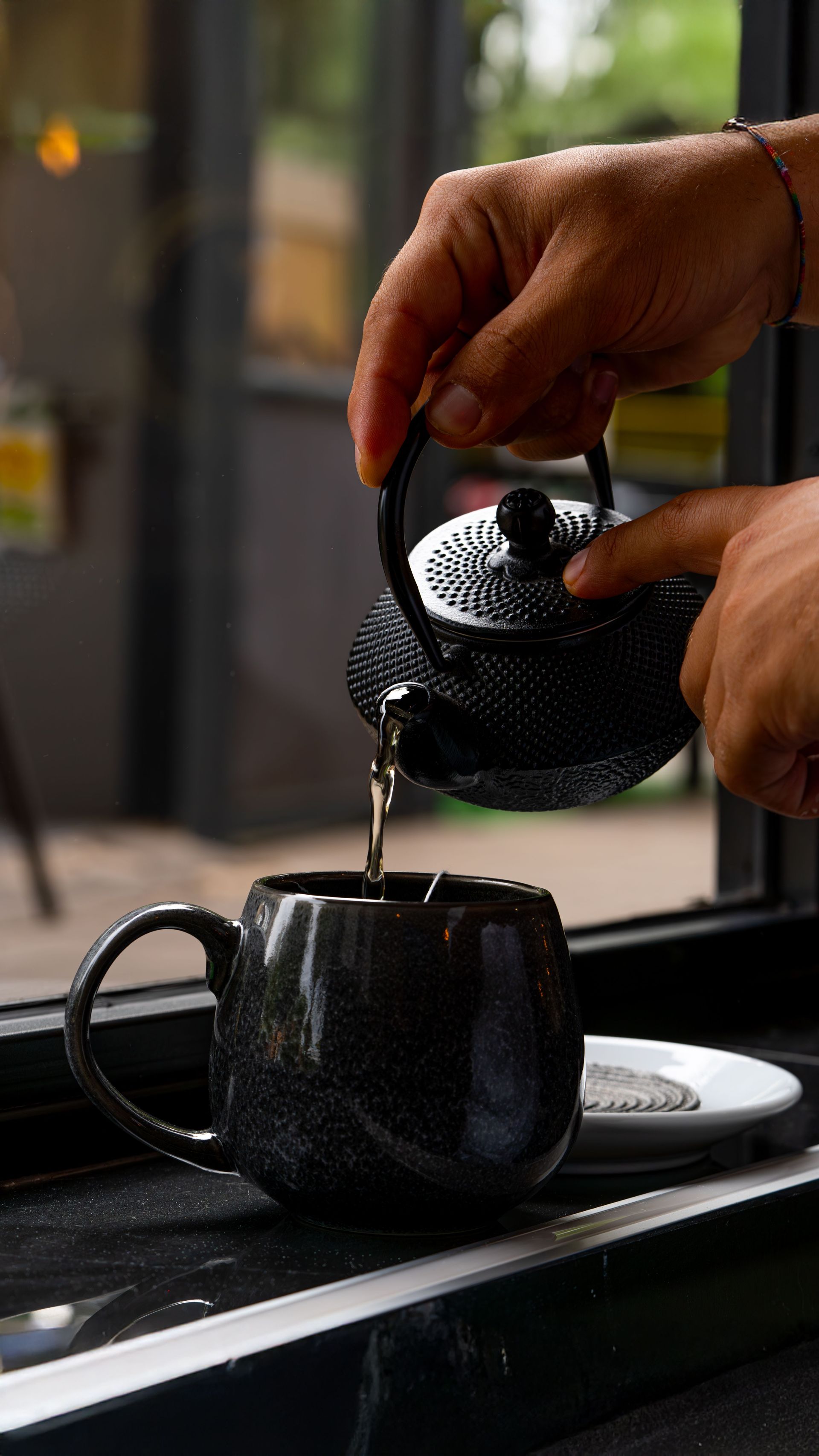 Des mains versent du thé d'une théière noire dans une tasse noire près d'une fenêtre.