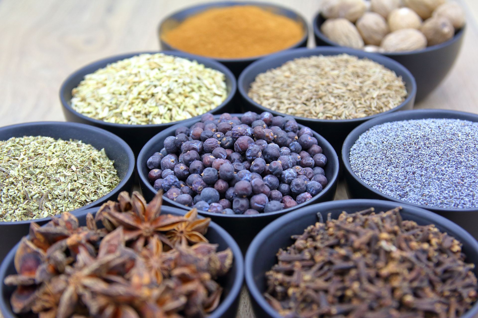 Assortiment d'épices et d'herbes aromatiques dans des bols noirs, comprenant notamment de l'anis étoilé, des grains de poivre et des graines séchées.