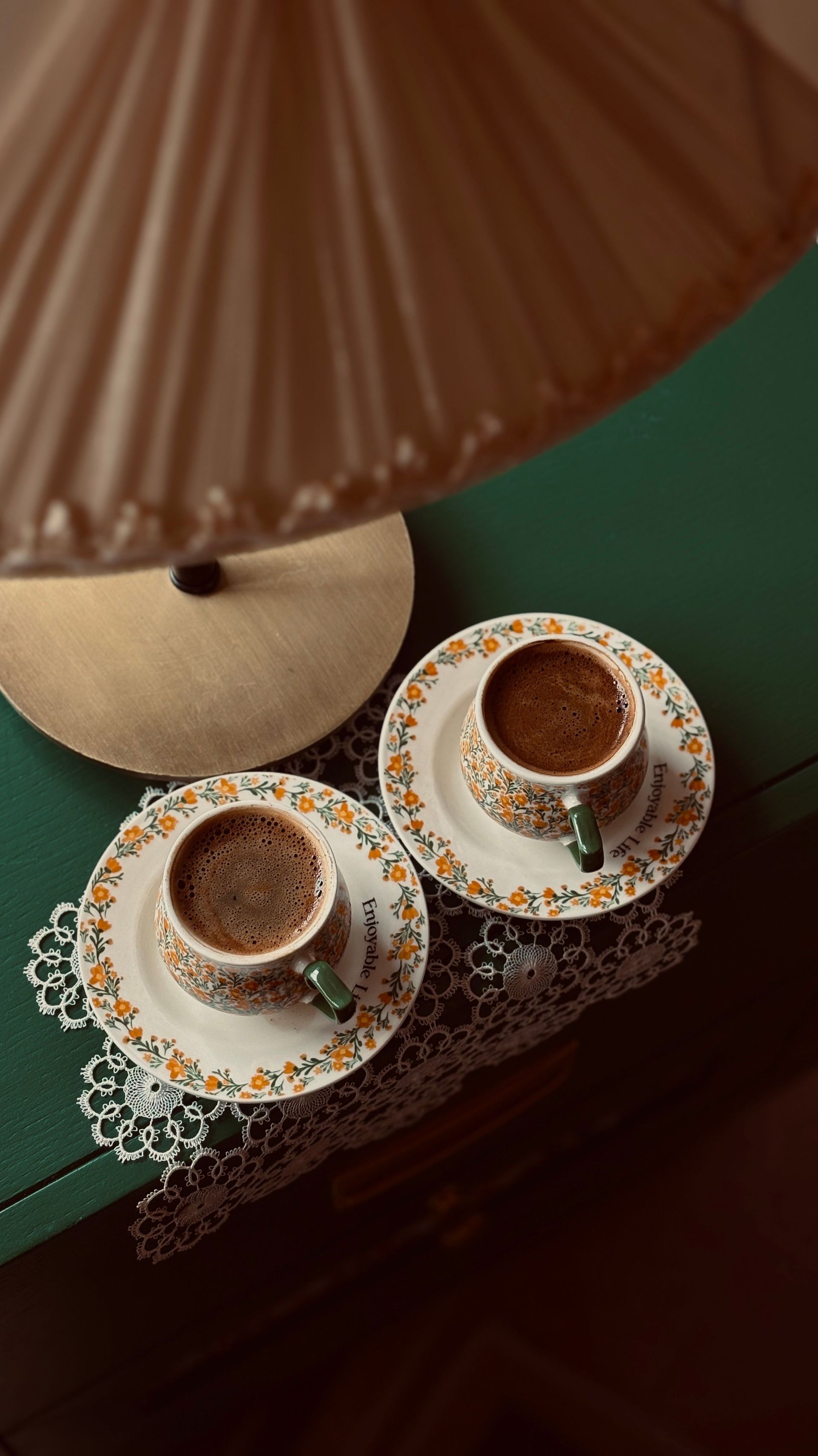 Deux tasses à thé ornées sur leurs soucoupes assorties, à côté d'une lampe beige sur une table vert foncé.