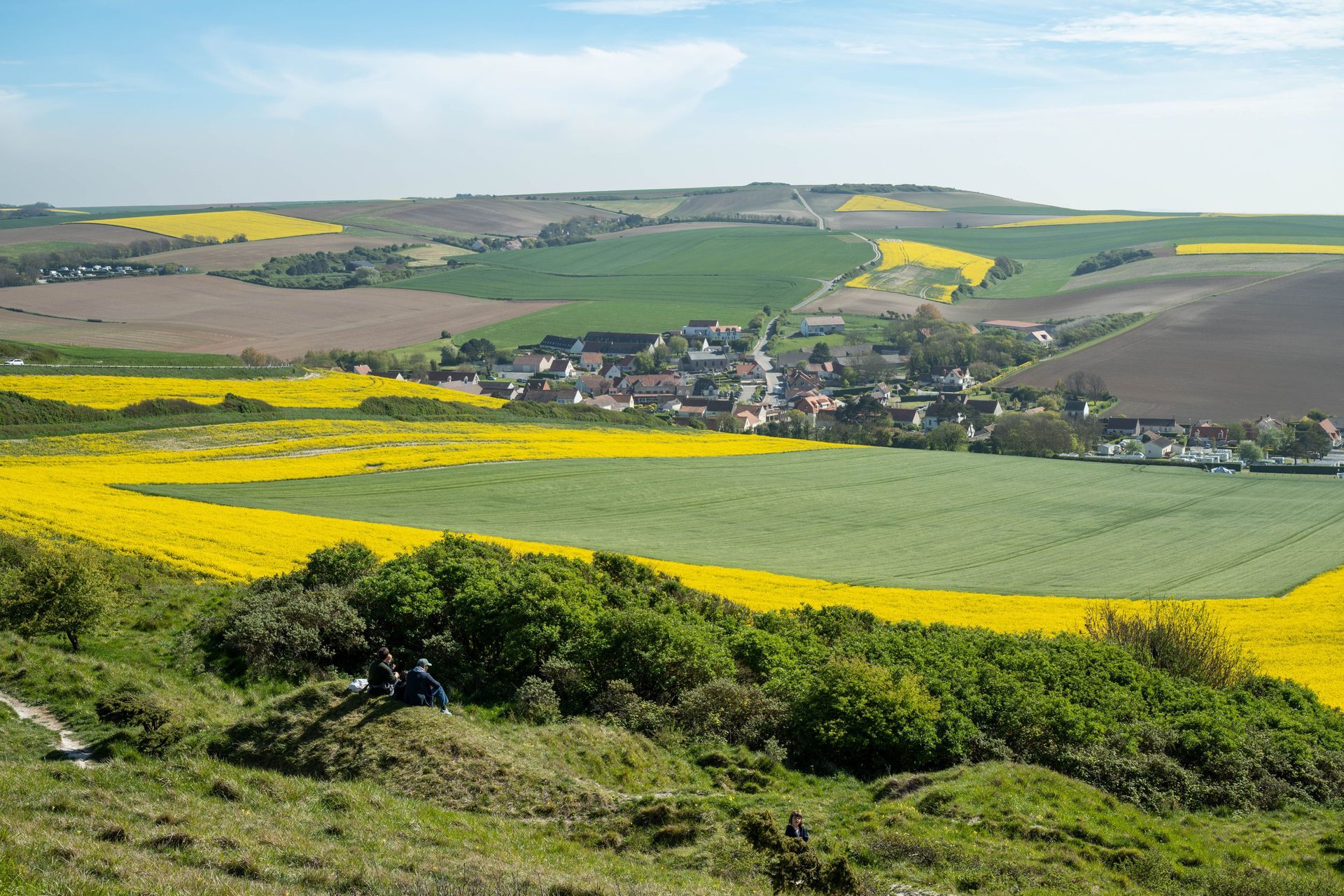 Des terres agricoles vallonnées, vertes et jaunes, avec un petit village dans une vallée sous un ciel bleu.