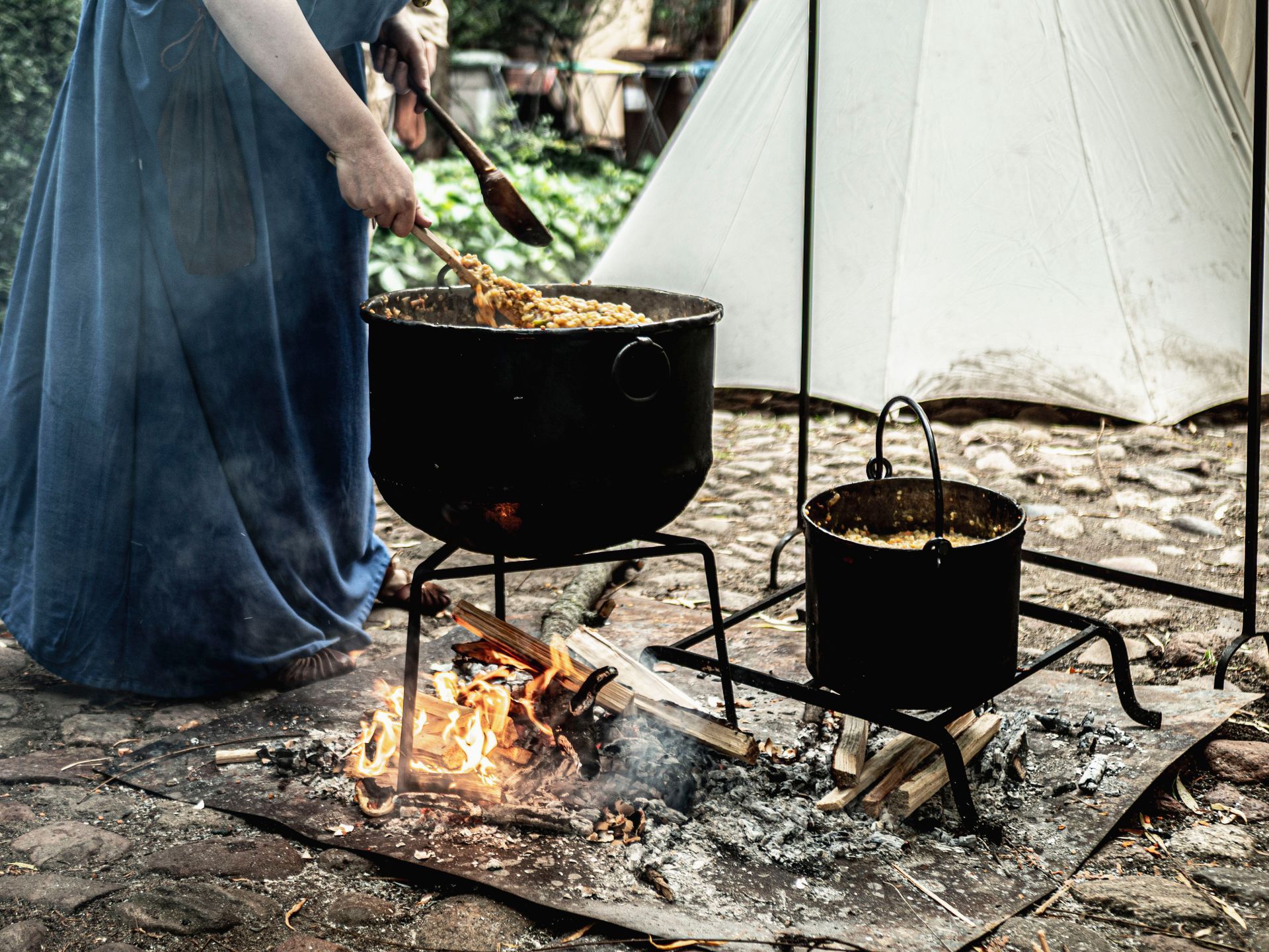 Une personne cuisine sur un feu de bois à côté d'une tente blanche, avec deux casseroles noires sur trépieds.