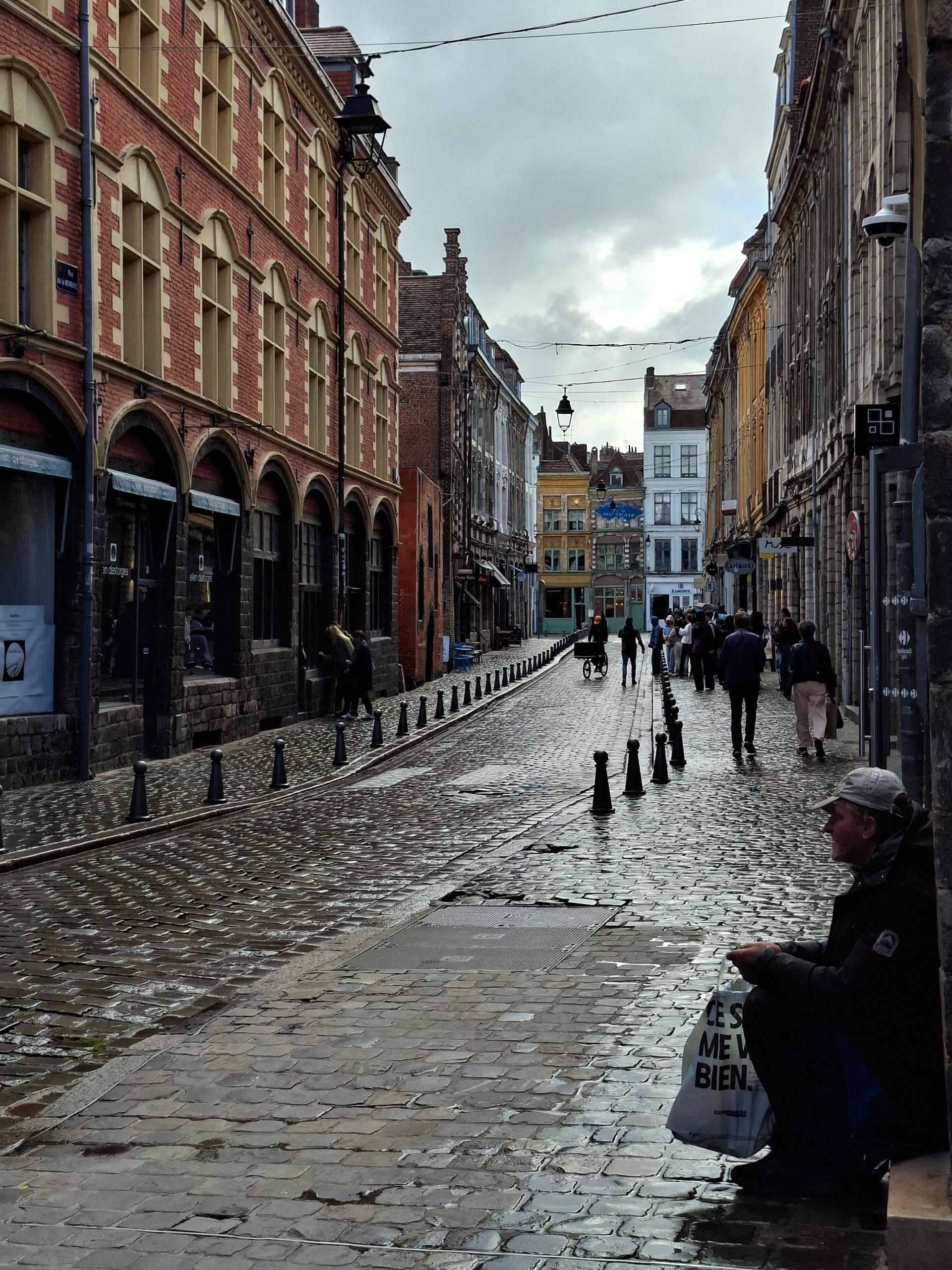 Rue pavée bordée de vieux bâtiments en briques par une journée pluvieuse, avec quelques piétons et une personne assise.