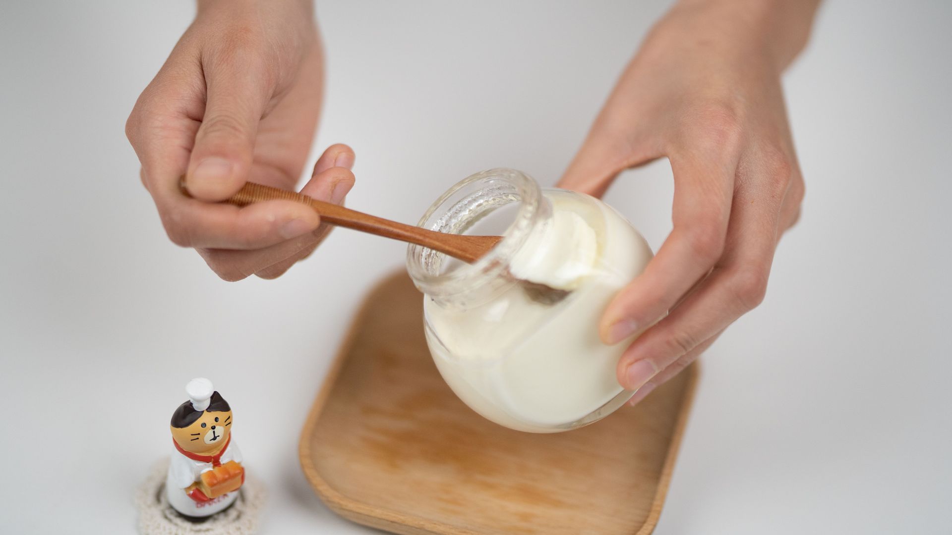 Des mains remuent de la crème blanche dans un pot en verre posé sur un plateau, avec une petite figurine à proximité.