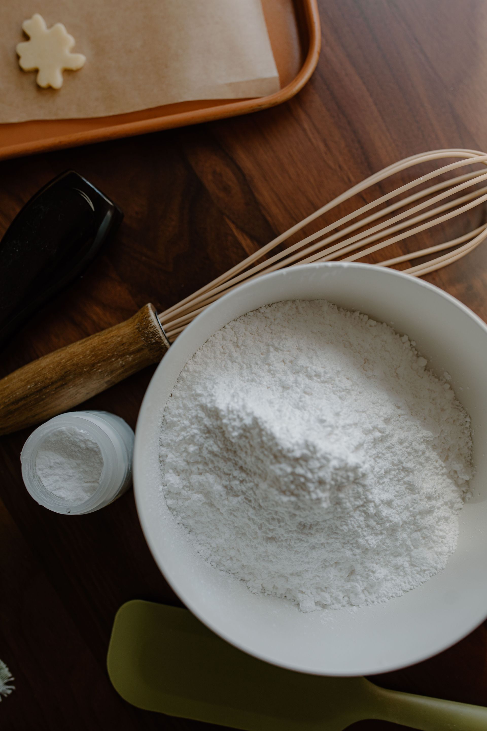 Préparation du matériel de pâtisserie : farine dans un bol, fouet, cuillères doseuses et planche à pâtisserie sur une table en bois.