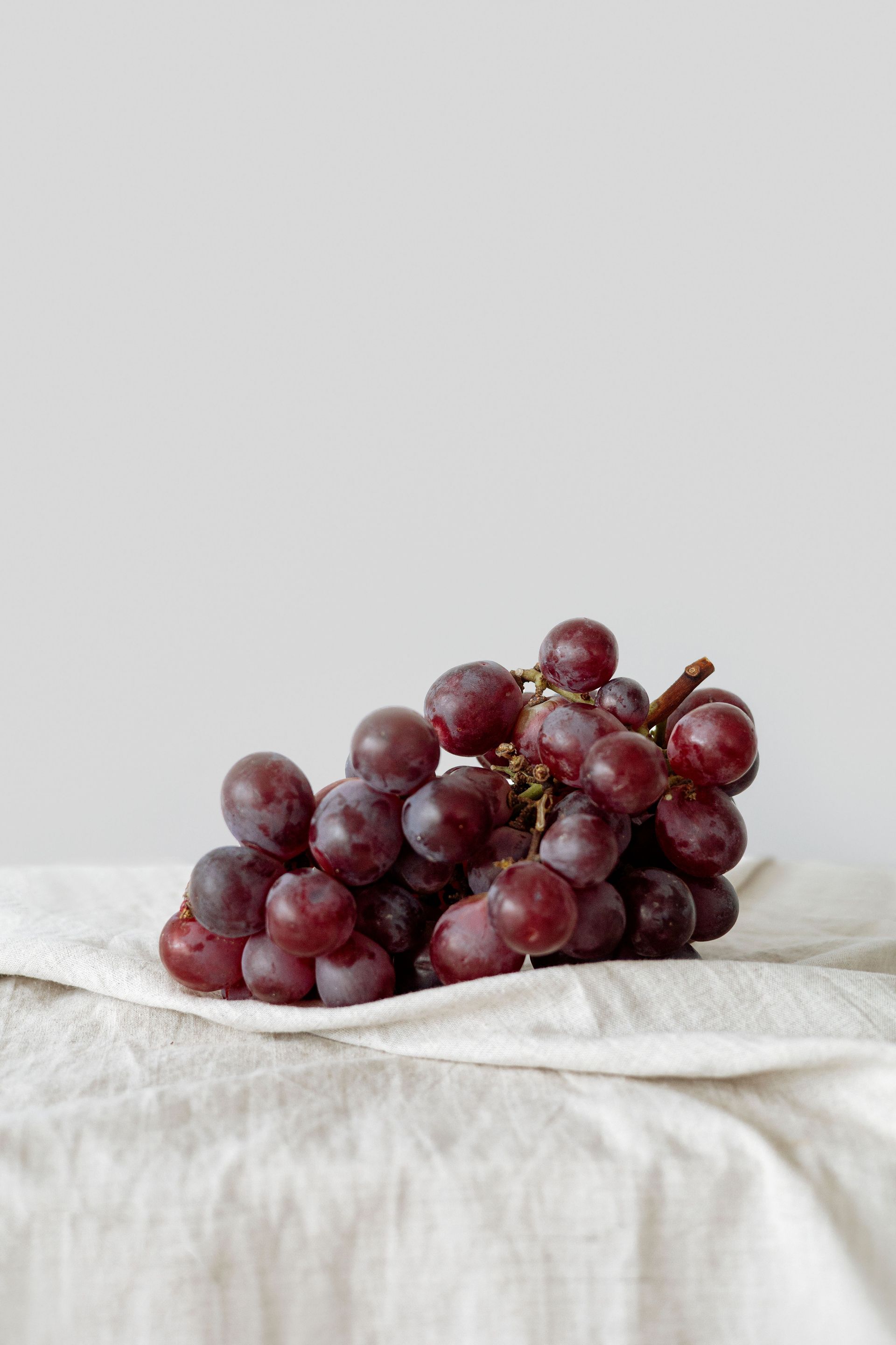 Une grappe de raisins rouges posée sur un tissu blanc devant un fond clair uni