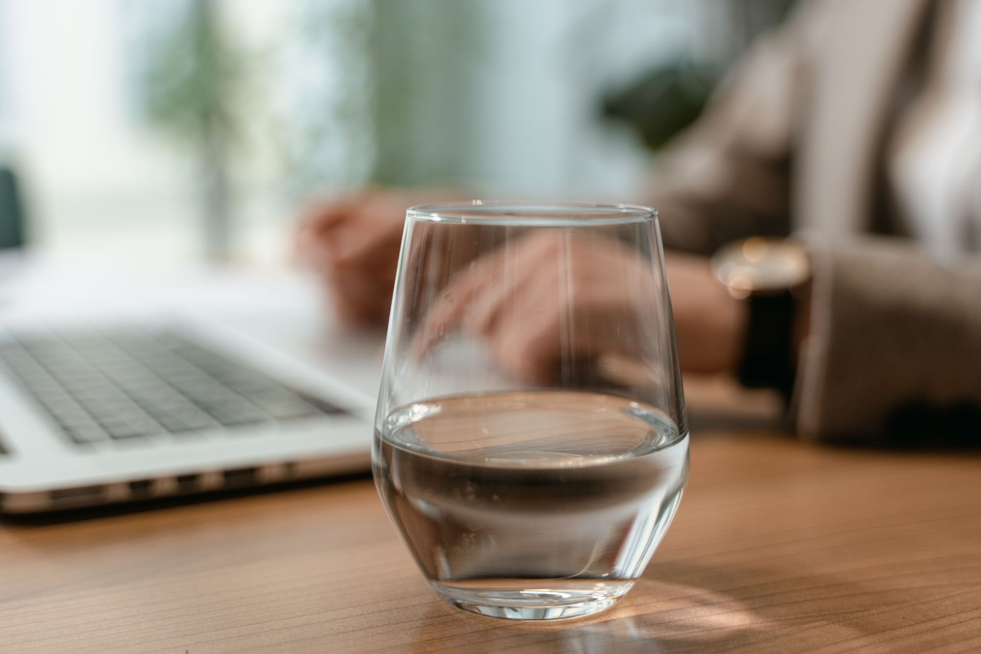 Un verre d'eau sur un bureau en bois à côté d'un ordinateur portable, avec une personne floue en arrière-plan.