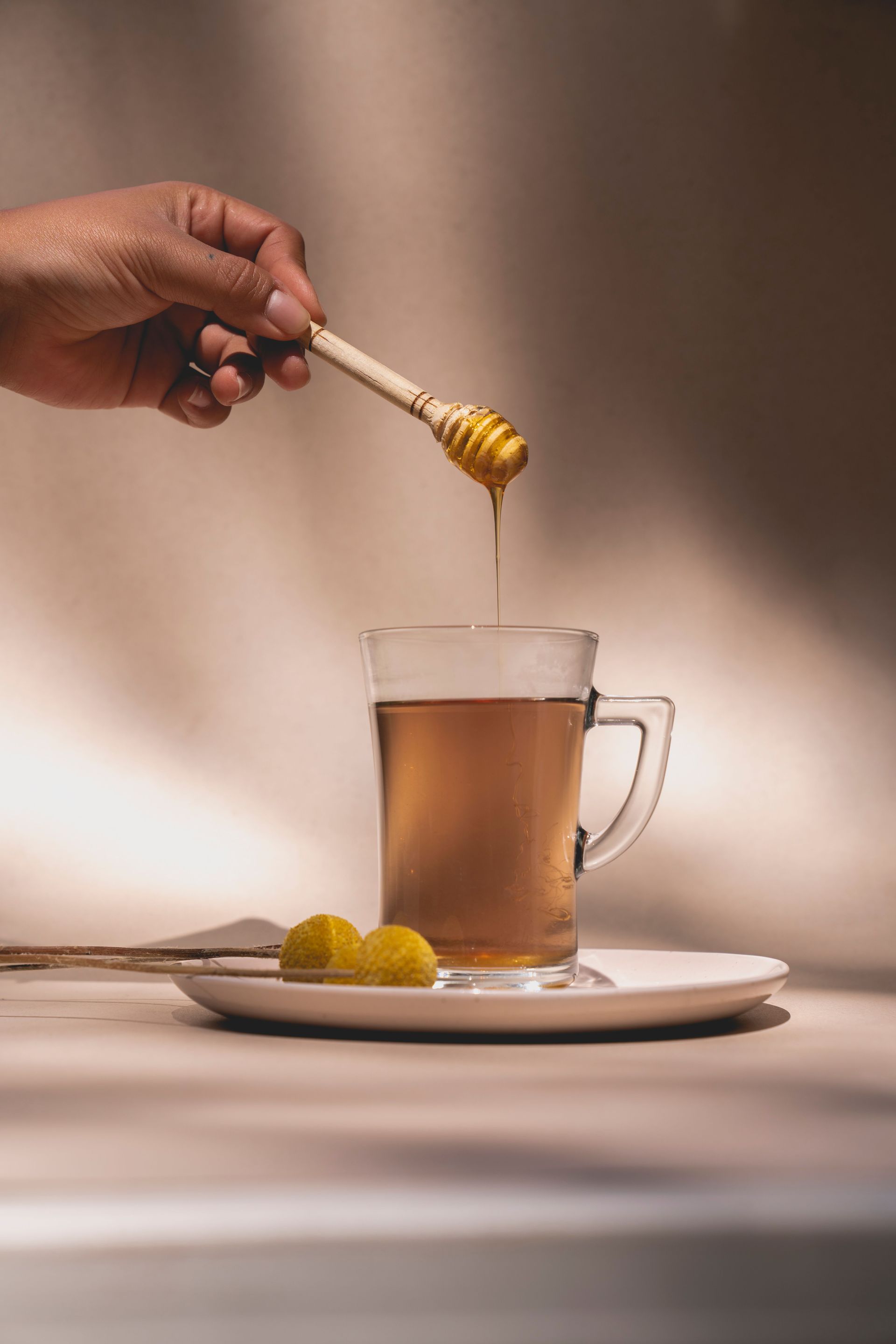 Une main verse du miel dans une tasse de thé en verre posée sur une soucoupe garnie de tranches de citron.