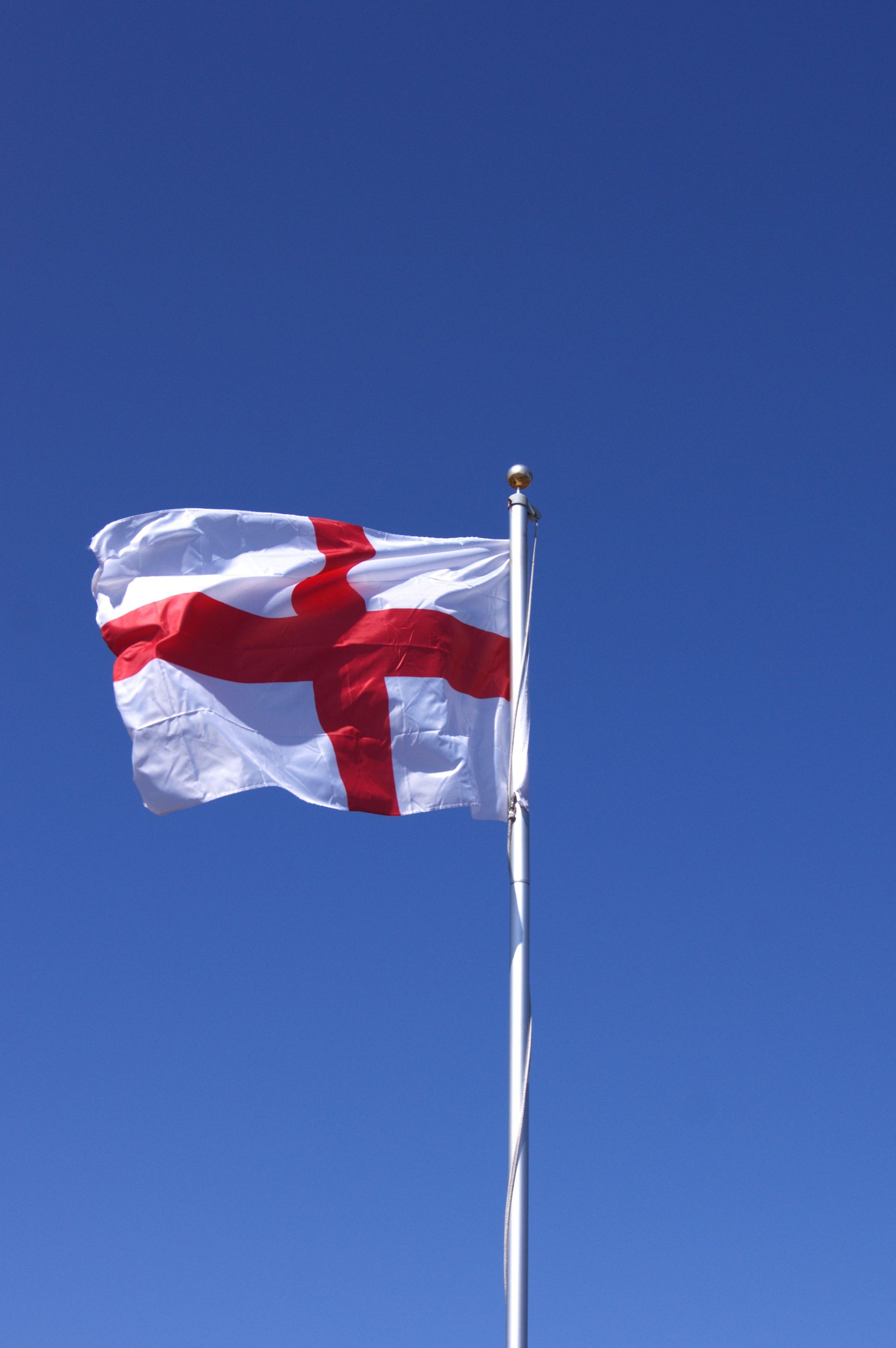 Un drapeau blanc à croix rouge flotte au sommet d'un mât sur un ciel bleu clair.