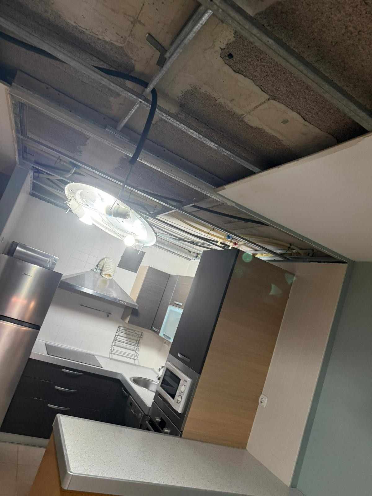 Interior de cocina inclinado con techo a la vista, lámpara colgante, armarios blancos y una puerta abierta.