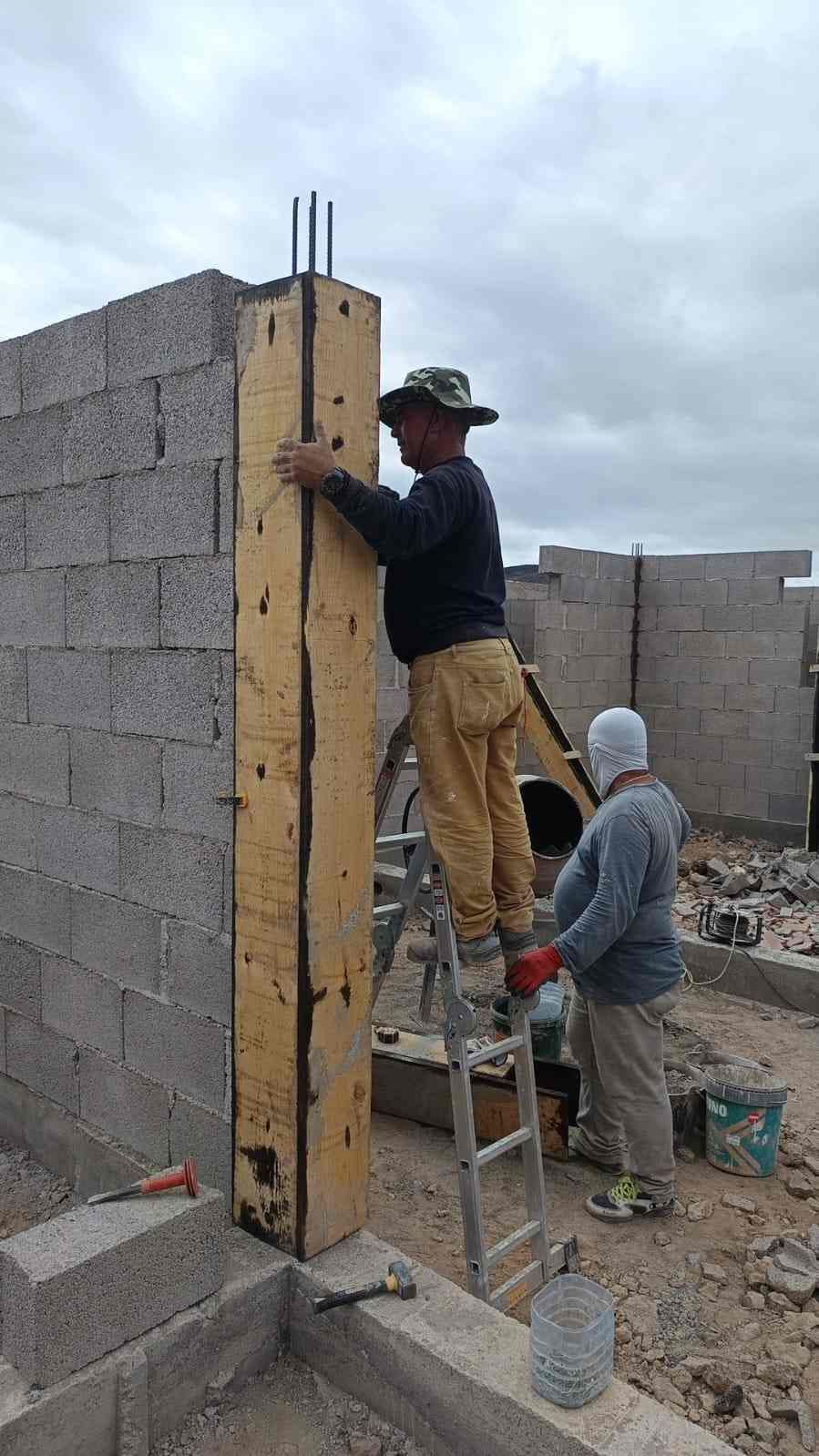Dos trabajadores construyen un muro de bloques de hormigón; uno de ellos está subido a una escalera.