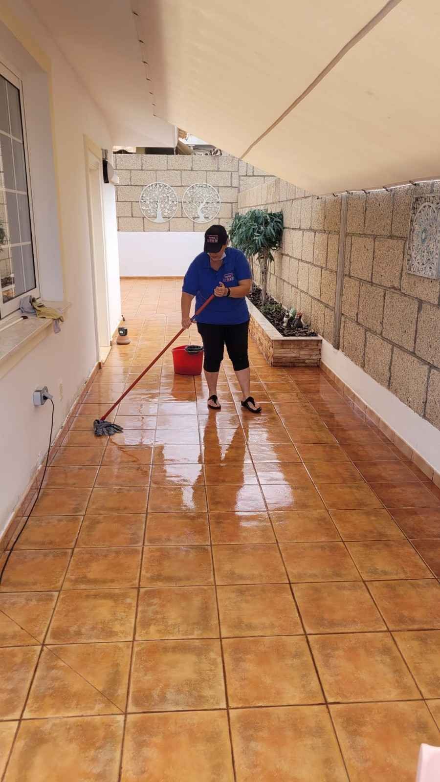 Persona que friega un pasillo embaldosado con un cubo y una escoba rojos, vestida con una camisa azul.