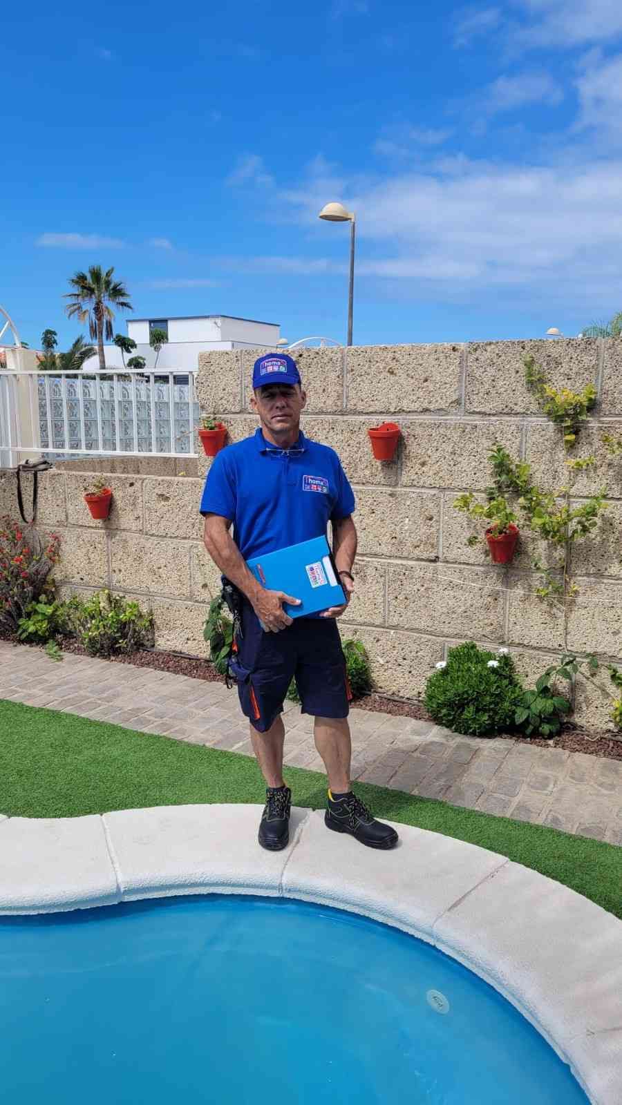 Un hombre de pie junto a un estanque sosteniendo una carpeta azul, con un muro de piedra, plantas y cielo azul detrás.