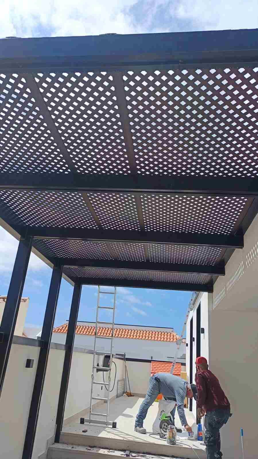 Trabajadores instalando materiales bajo una pérgola de celosía negra junto a un edificio blanco en un día soleado.