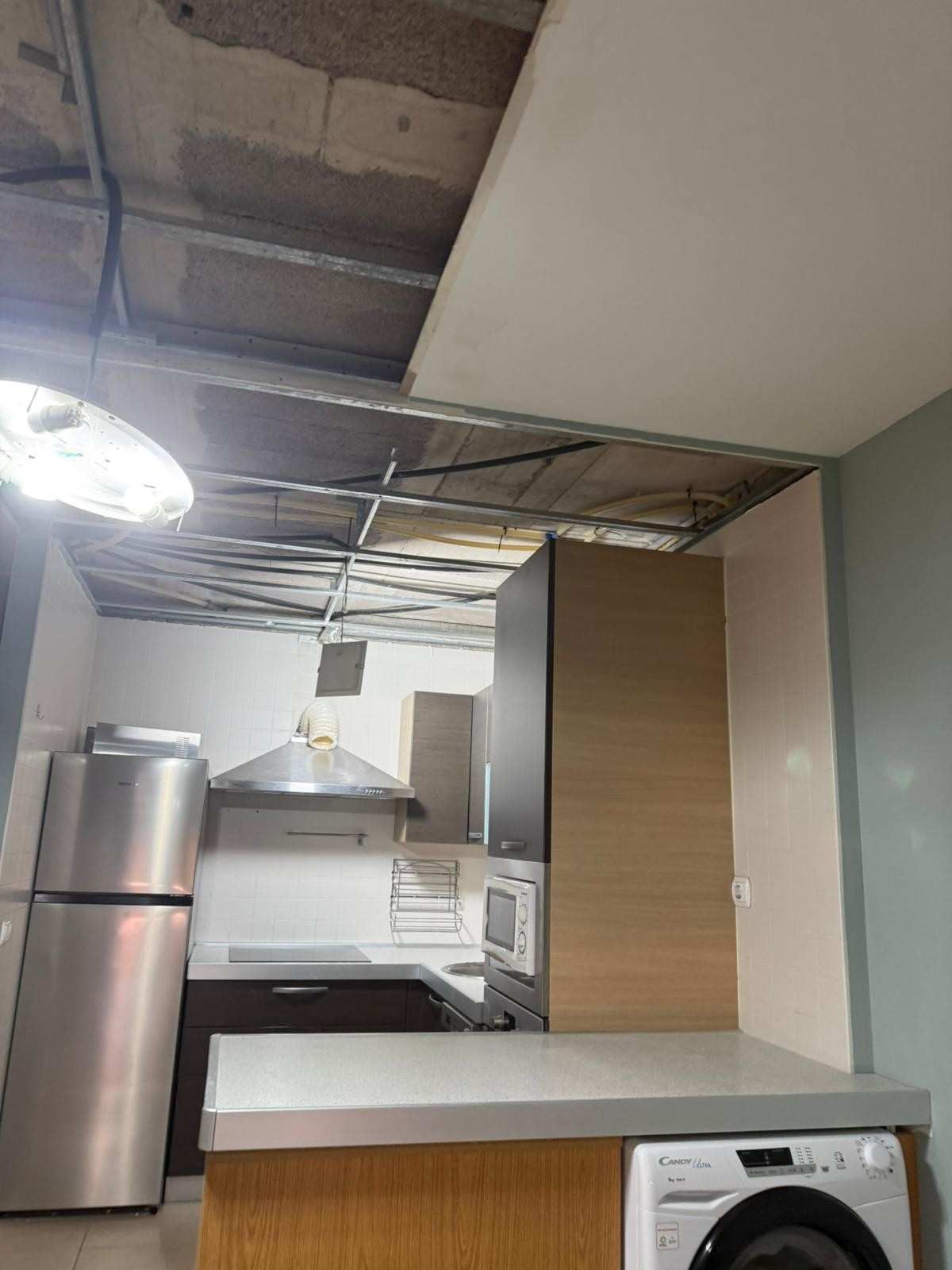 Cocina parcialmente sin terminar con techo a la vista, nevera de acero inoxidable, armarios blancos y lavadora a la derecha.