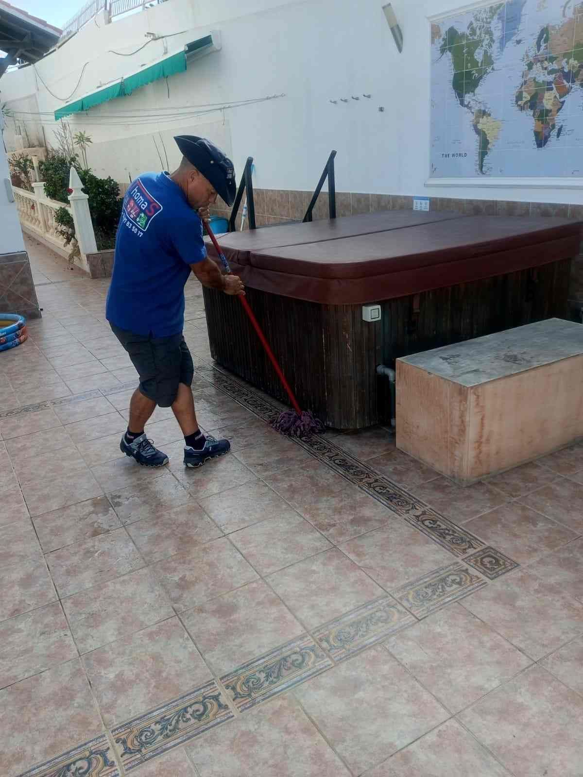 Una persona con camisa azul está fregando un patio de baldosas junto a una bañera de hidromasaje elevada y un mapa mural.