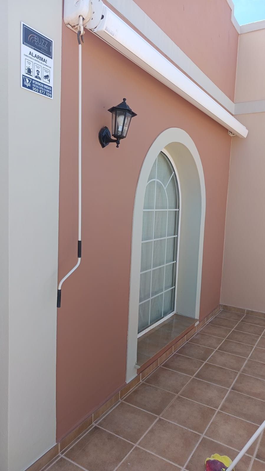 Pared exterior rosa con puerta de cristal arqueada, lámpara de pared y pasillo embaldosado.