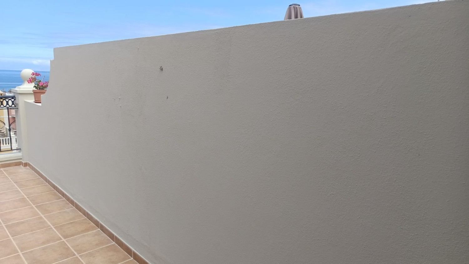 Gran pared beige junto a un camino de baldosas, con el océano visible a la izquierda bajo un cielo despejado.